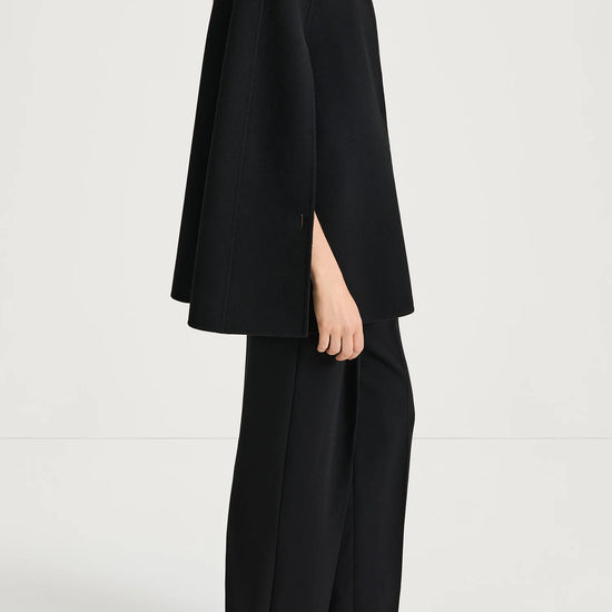 A model shown in profile wearing a black high-neck cape-style top with wide sleeves and matching black wide-leg trousers, photographed against a clean light gray studio background.