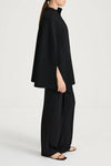 A model shown in profile wearing a black high-neck cape-style top with wide sleeves and matching black wide-leg trousers, photographed against a clean light gray studio background.