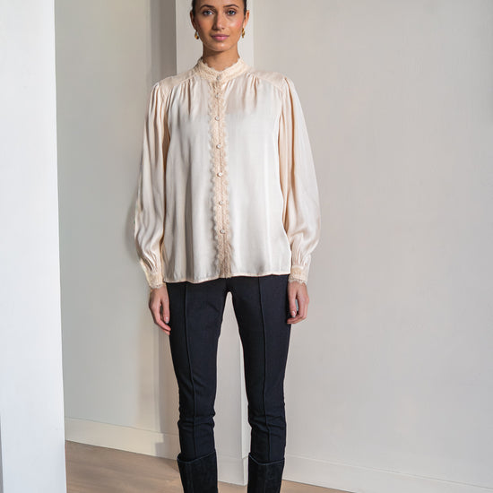 A woman modeling a cream-colored long-sleeve blouse with a ruffled lace front and high collar, paired with black pants and black knee-high boots, standing in a minimalist room with white walls and light wood floors.