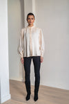 A woman modeling a cream-colored long-sleeve blouse with a ruffled lace front and high collar, paired with black pants and black knee-high boots, standing in a minimalist room with white walls and light wood floors.