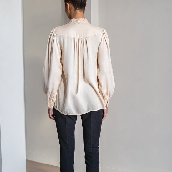 A woman viewed from behind wearing a cream-colored long-sleeve blouse with gathered detailing and dark pants, standing in a minimalist room with white walls and light wood flooring.