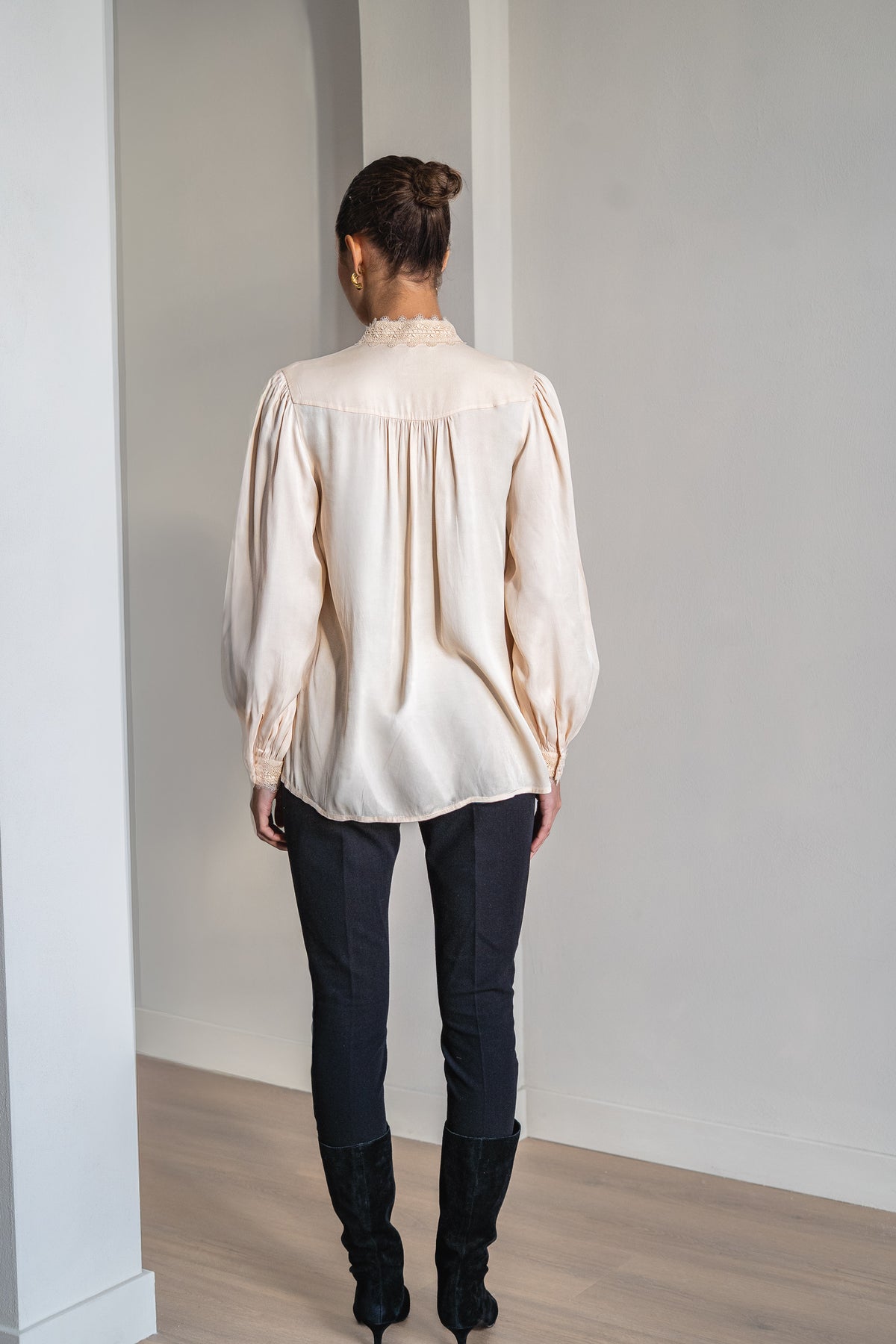 A woman viewed from behind wearing a cream-colored long-sleeve blouse with gathered detailing and dark pants, standing in a minimalist room with white walls and light wood flooring.