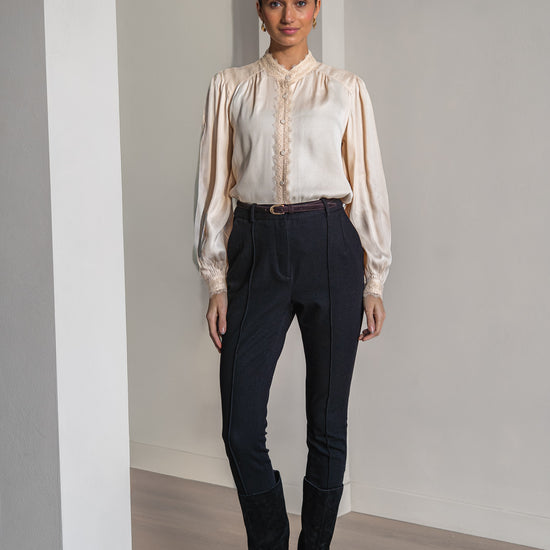 A woman modeling a cream satin blouse with lace detailing and dark navy trousers, standing in a minimalist room with light wood floors and white walls.