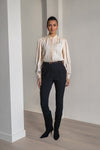 A woman modeling a cream satin blouse with lace detailing and dark navy trousers, standing in a minimalist room with light wood floors and white walls.