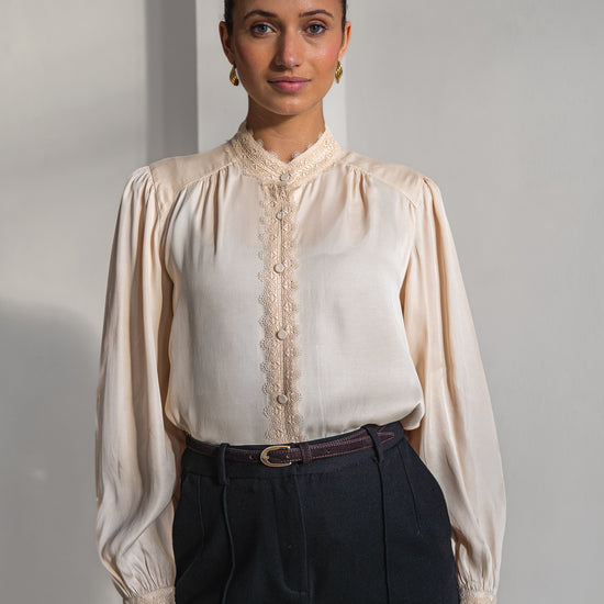 A woman modeling a cream-colored long-sleeve blouse with lace trim and a high collar, paired with dark trousers, set against a minimalist light gray background.
