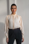 A woman modeling a cream-colored long-sleeve blouse with lace trim and a high collar, paired with dark trousers, set against a minimalist light gray background.