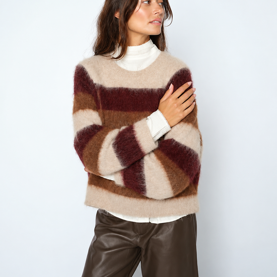 A woman wearing a fuzzy striped sweater in beige, brown, and burgundy, layered over a white turtleneck, paired with dark brown faux-leather pants, against a light gray studio background.