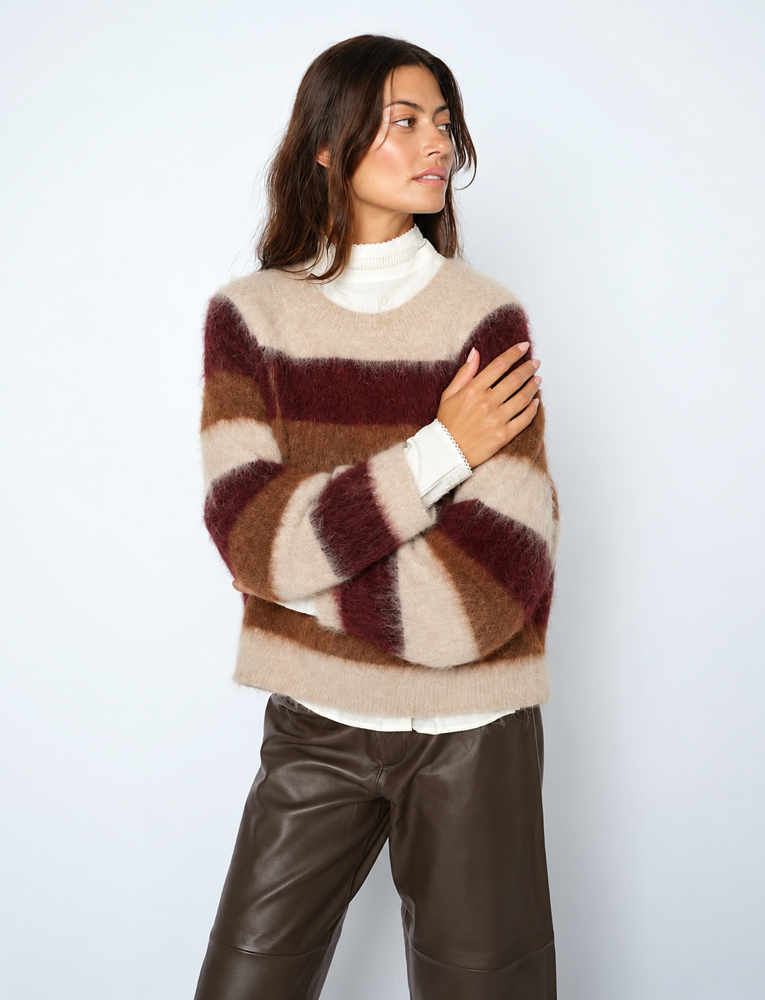 A woman wearing a fuzzy striped sweater in beige, brown, and burgundy, layered over a white turtleneck, paired with dark brown faux-leather pants, against a light gray studio background.