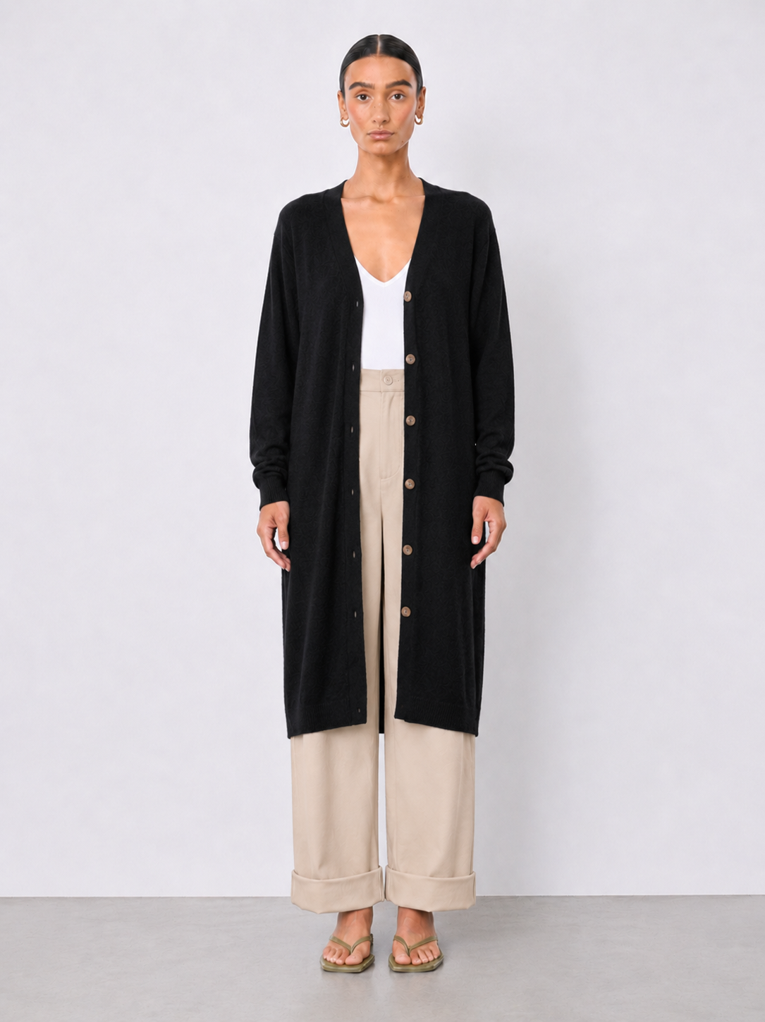 A model stands against a light gray studio background wearing a long black cardigan over a white top and beige wide-leg cropped pants.
