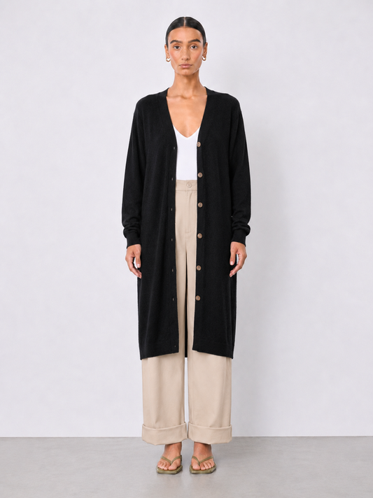 A model stands against a light gray studio background wearing a long black cardigan over a white top and beige wide-leg cropped pants.