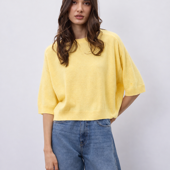 A woman modeling a bright yellow short-sleeve knit top with relaxed, oversized fit and blue wide-leg jeans against a light gray studio background.