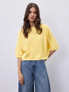 A woman modeling a bright yellow short-sleeve knit top with relaxed, oversized fit and blue wide-leg jeans against a light gray studio background.