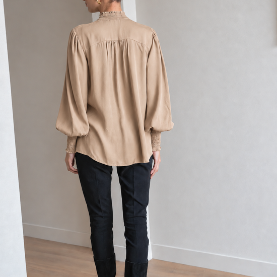 A woman viewed from behind wearing a beige long-sleeve blouse with a gathered, pleated back and puff sleeves, paired with black pants and black knee-high boots in a minimalist room with light gray walls and a wood floor.