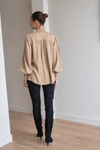 A woman viewed from behind wearing a beige long-sleeve blouse with a gathered, pleated back and puff sleeves, paired with black pants and black knee-high boots in a minimalist room with light gray walls and a wood floor.
