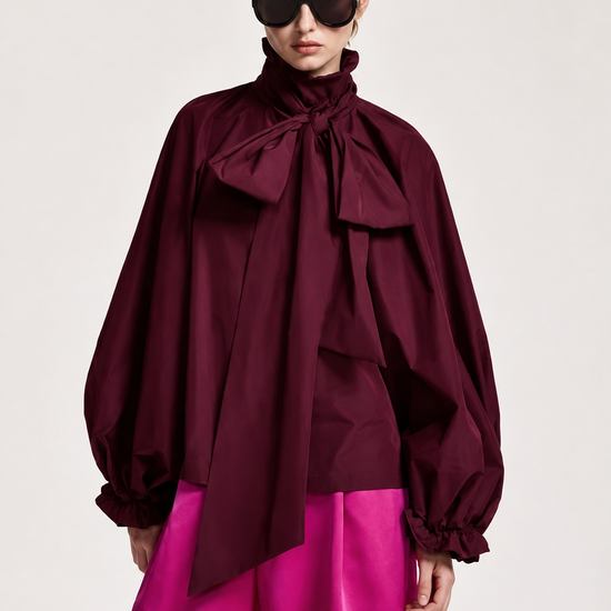 A model wearing a dark burgundy high-neck blouse with dramatic balloon sleeves and a large front bow, paired with bright fuchsia pants, against a plain light gray studio background.