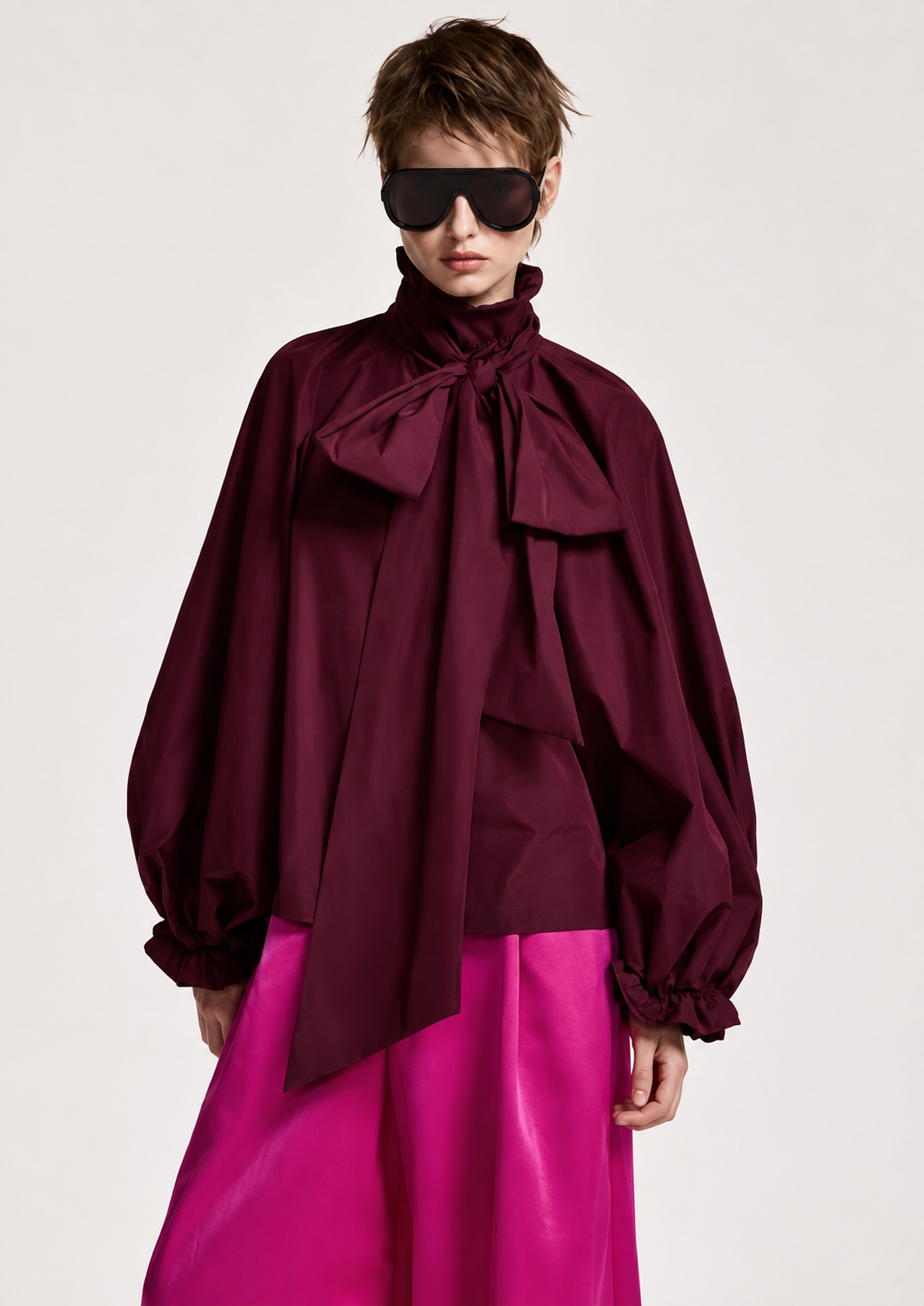 A model wearing a dark burgundy high-neck blouse with dramatic balloon sleeves and a large front bow, paired with bright fuchsia pants, against a plain light gray studio background.