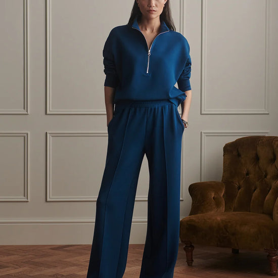 A model stands in a classic room with paneled walls and a wooden floor, wearing a matching navy blue two-piece set consisting of a half-zip long-sleeve top and wide-leg trousers. A brown velvet armchair is visible on the right.