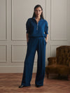 A model stands in a classic room with paneled walls and a wooden floor, wearing a matching navy blue two-piece set consisting of a half-zip long-sleeve top and wide-leg trousers. A brown velvet armchair is visible on the right.