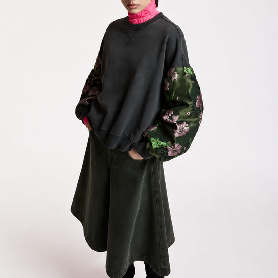 A model wearing a black oversized sweatshirt with green floral patterned balloon sleeves, a dark olive green skirt, and black pointed-toe boots, standing against a plain light gray studio background.