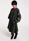 A model wearing a black oversized sweatshirt with green floral patterned balloon sleeves, a dark olive green skirt, and black pointed-toe boots, standing against a plain light gray studio background.