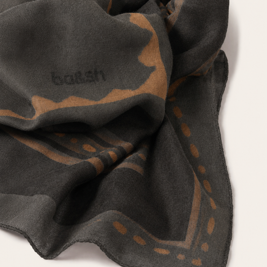 A dark charcoal gray scarf with a warm brown abstract pattern and the brand name 'ba&sh' woven into it, shown on a light beige background.