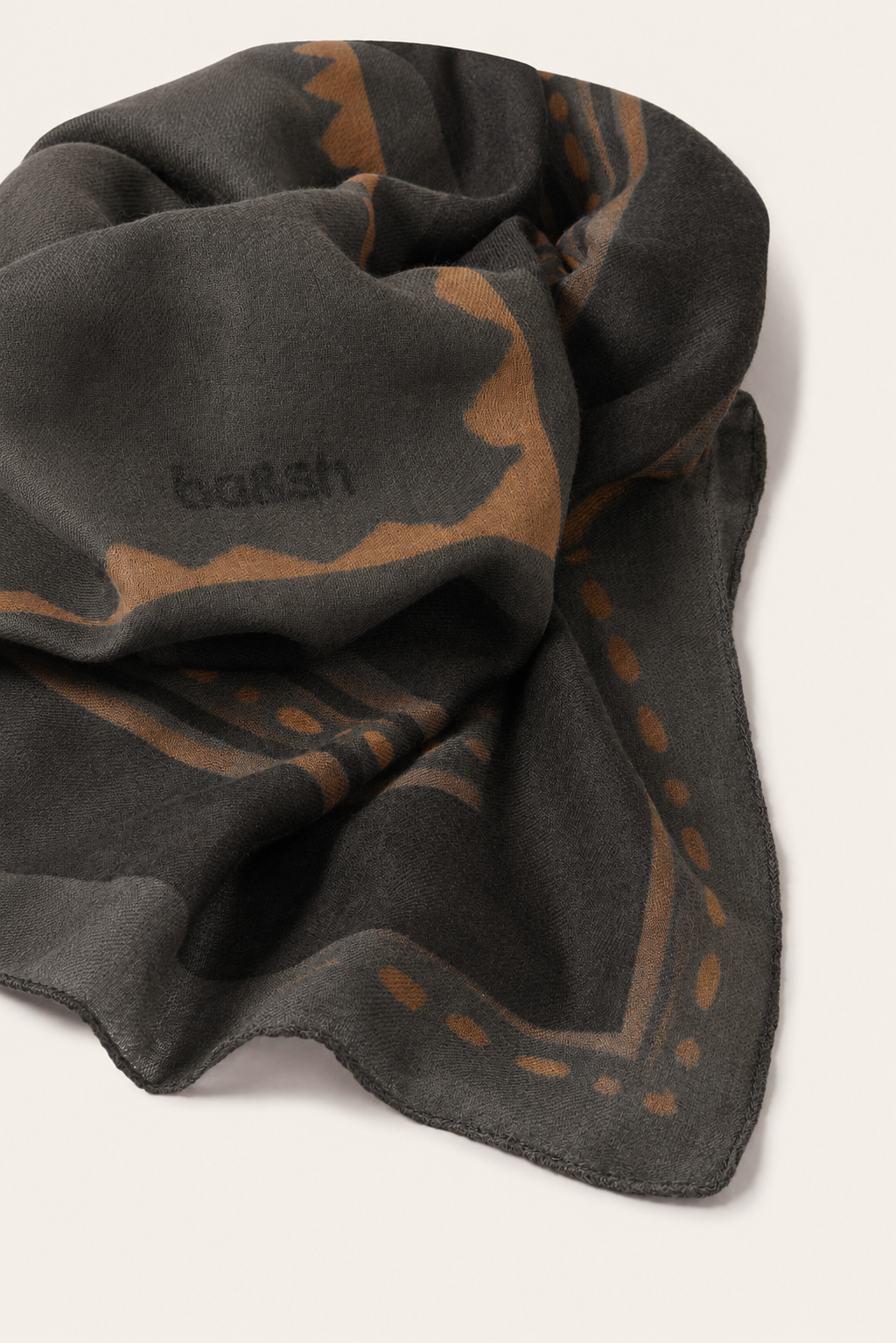 A dark charcoal gray scarf with a warm brown abstract pattern and the brand name 'ba&sh' woven into it, shown on a light beige background.