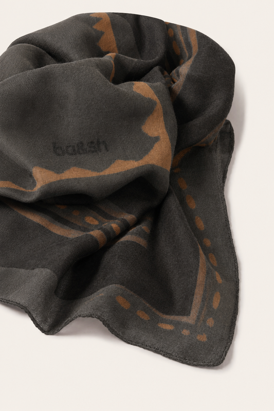 A dark charcoal gray scarf with a warm brown abstract pattern and the brand name 'ba&sh' woven into it, shown on a light beige background.