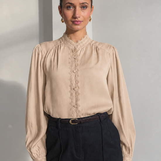 A woman modeling a beige long-sleeve blouse with lace trim and a buttoned high collar, paired with dark trousers, against a minimalist light gray wall.