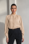 A woman modeling a beige long-sleeve blouse with lace trim and a buttoned high collar, paired with dark trousers, against a minimalist light gray wall.