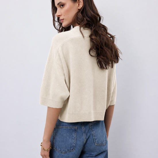A woman shown from the back wearing a cream-colored short-sleeve knit top and blue jeans against a light gray studio background.