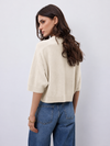 A woman shown from the back wearing a cream-colored short-sleeve knit top and blue jeans against a light gray studio background.