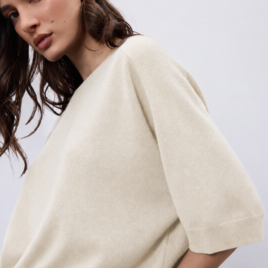 A close-up of a woman wearing a cream-colored knit sweater with elbow-length sleeves, shown against a plain light gray background.