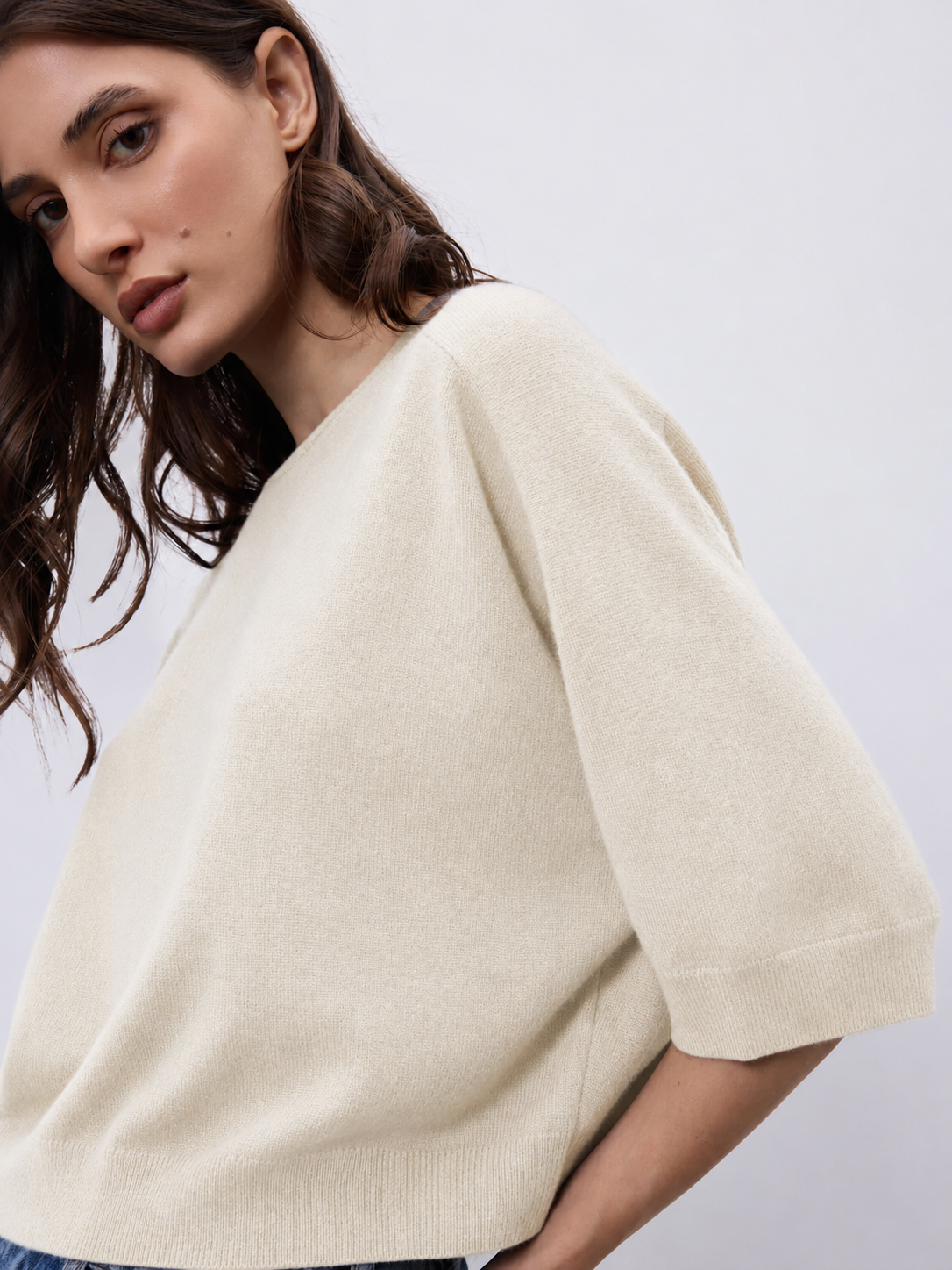A close-up of a woman wearing a cream-colored knit sweater with elbow-length sleeves, shown against a plain light gray background.