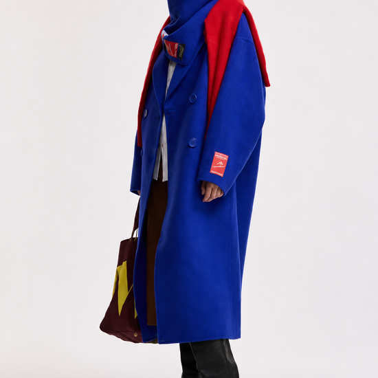 A model stands in a studio wearing a long bright blue coat over a red and navy layered top, with black knee-high boots and a patterned brown and yellow bag, against a plain light gray background.