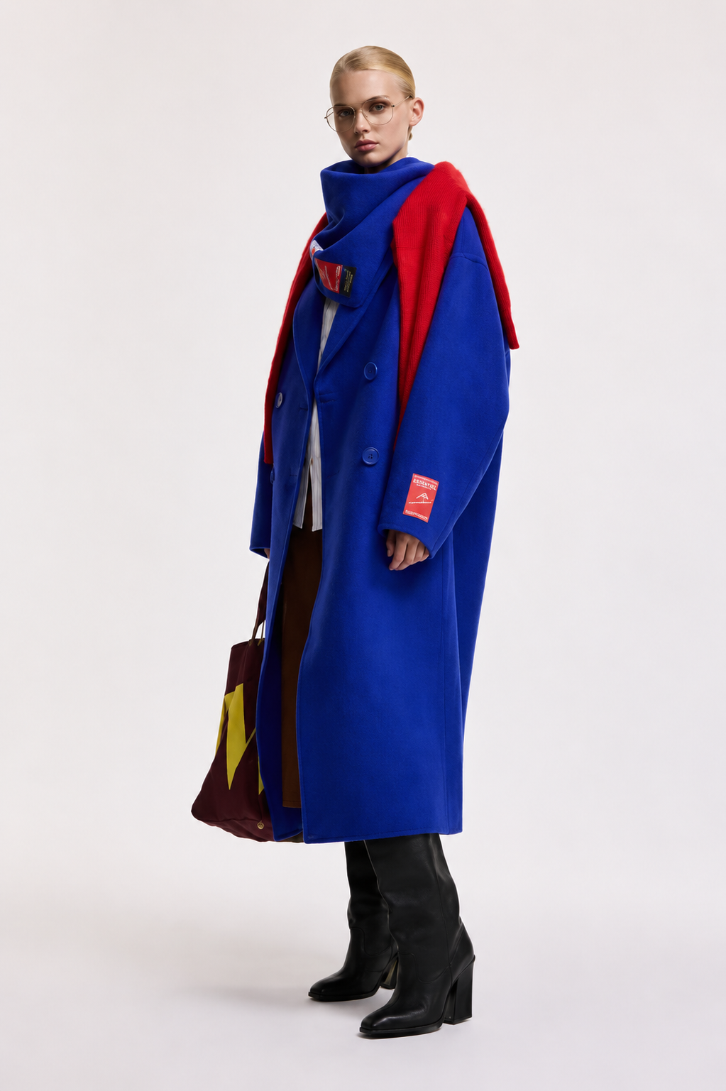 A model stands in a studio wearing a long bright blue coat over a red and navy layered top, with black knee-high boots and a patterned brown and yellow bag, against a plain light gray background.