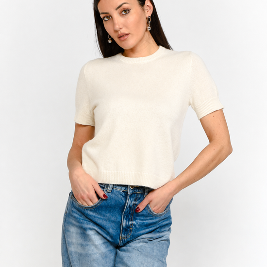 A woman modeling a short-sleeve cream knit top paired with blue jeans, standing against a plain light gray studio background.