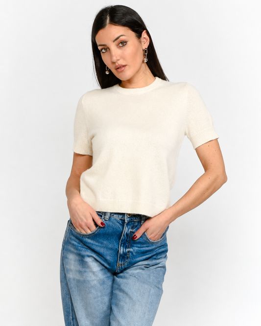 A woman modeling a short-sleeve cream knit top paired with blue jeans, standing against a plain light gray studio background.