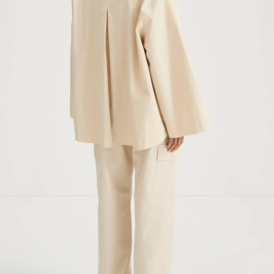A model shown from the back wearing a cream-colored, oversized two-piece suit consisting of a high-neck, wide-sleeved jacket and matching wide-leg trousers, paired with brown flat sandals against a plain white studio background.
