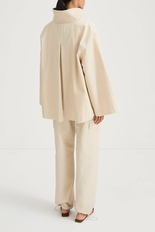 A model shown from the back wearing a cream-colored, oversized two-piece suit consisting of a high-neck, wide-sleeved jacket and matching wide-leg trousers, paired with brown flat sandals against a plain white studio background.