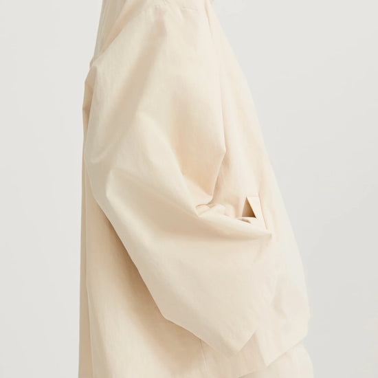 A side-profile view of a model wearing a cream-colored, oversized high-collar jacket and matching trousers against a plain light gray studio background.