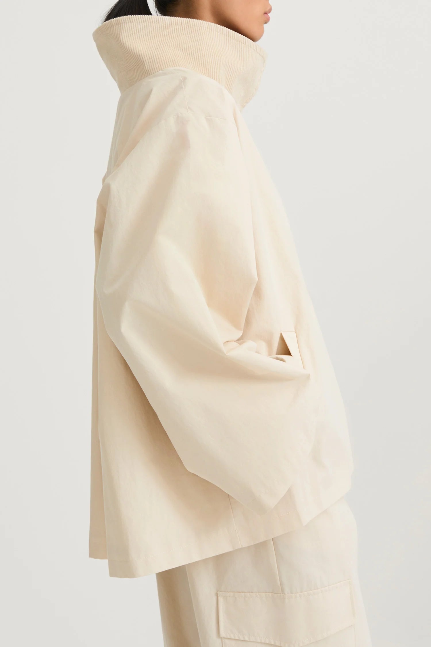A side-profile view of a model wearing a cream-colored, oversized high-collar jacket and matching trousers against a plain light gray studio background.