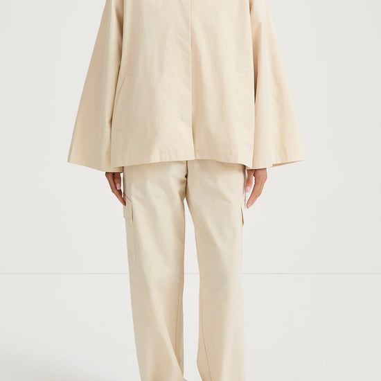 A model wearing a light beige high-collar jacket with wide bell sleeves and matching wide-leg trousers, paired with brown strappy sandals, shot against a plain white studio background.