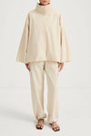 A model wearing a light beige high-collar jacket with wide bell sleeves and matching wide-leg trousers, paired with brown strappy sandals, shot against a plain white studio background.
