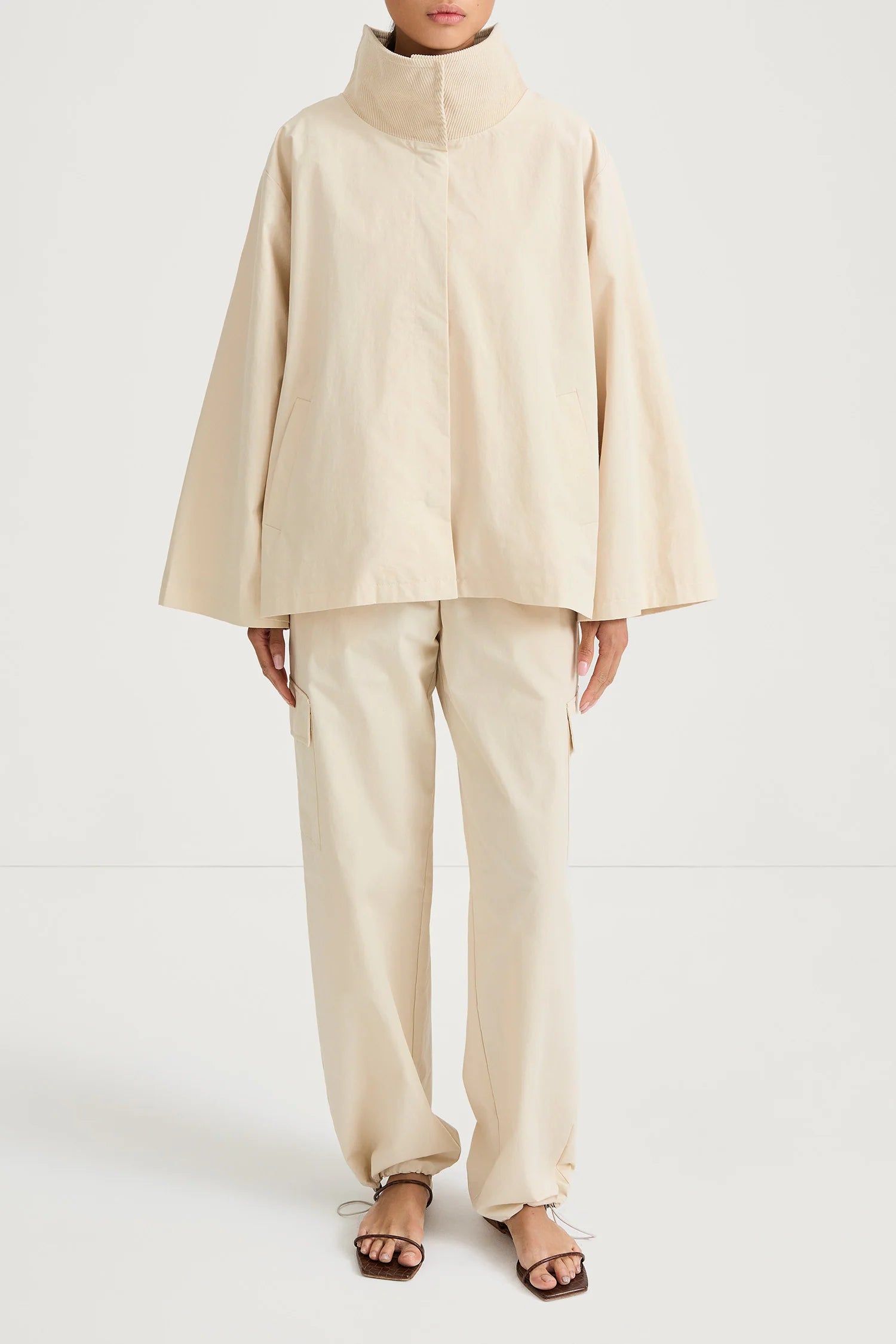 A model wearing a light beige high-collar jacket with wide bell sleeves and matching wide-leg trousers, paired with brown strappy sandals, shot against a plain white studio background.