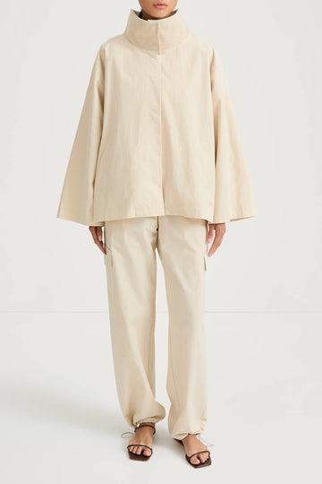 A model wearing a light beige high-collar jacket with wide bell sleeves and matching wide-leg trousers, paired with brown strappy sandals, shot against a plain white studio background.