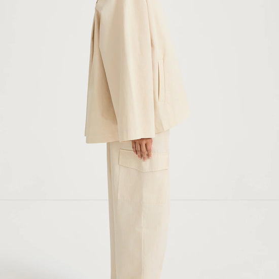 A model shown from the side wearing a cream-colored oversized jacket and matching wide-leg trousers, paired with brown leather sandals, against a plain white studio background.