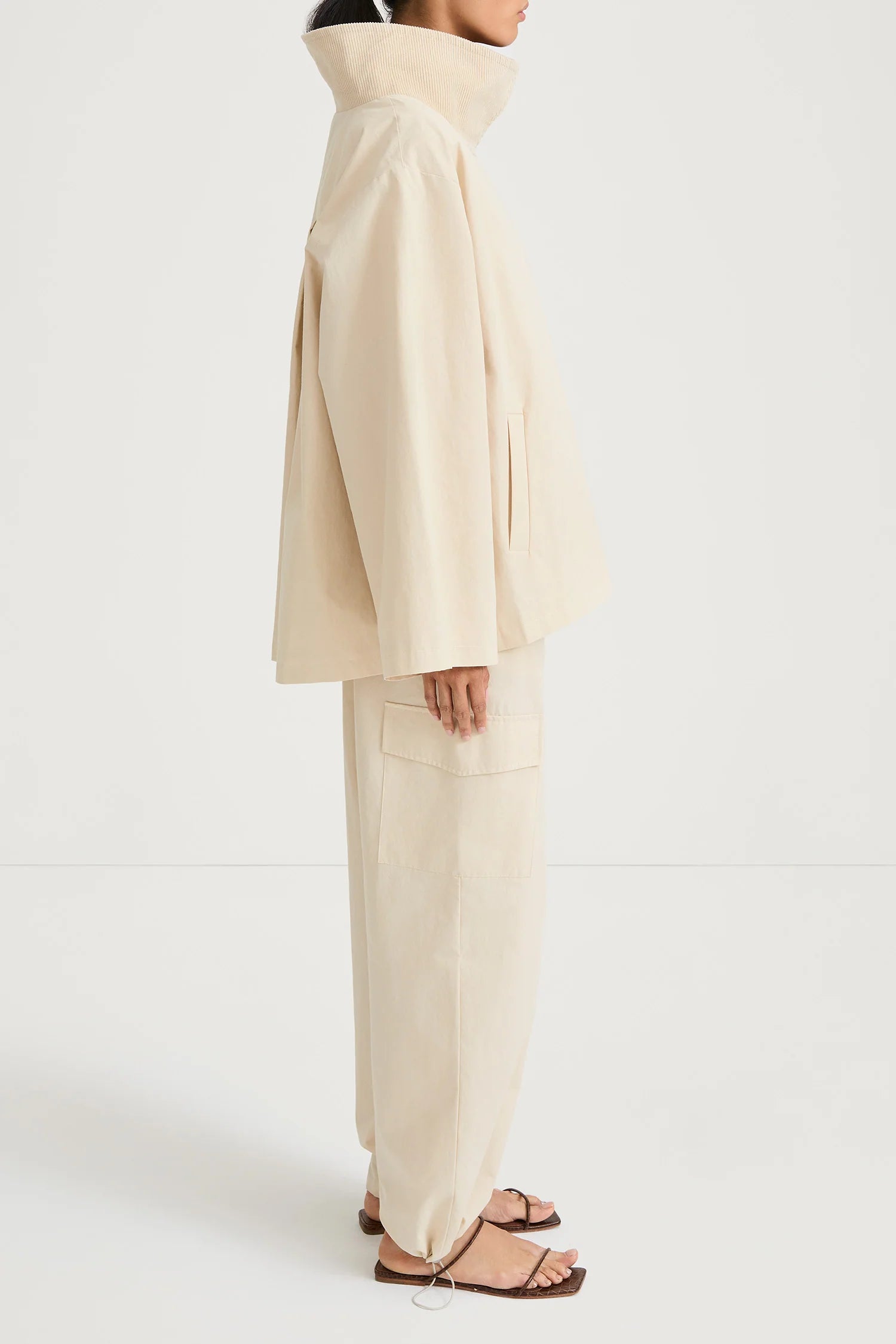 A model shown from the side wearing a cream-colored oversized jacket and matching wide-leg trousers, paired with brown leather sandals, against a plain white studio background.