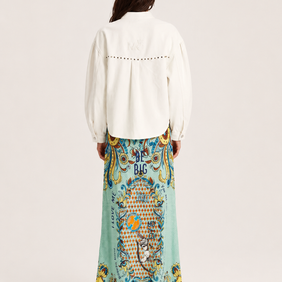 A woman shown from the back wearing a white long-sleeve blouse with decorative stitching and a long, wide skirt featuring a colorful baroque-style print in turquoise, gold, and blue.