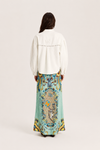 A woman shown from the back wearing a white long-sleeve blouse with decorative stitching and a long, wide skirt featuring a colorful baroque-style print in turquoise, gold, and blue.