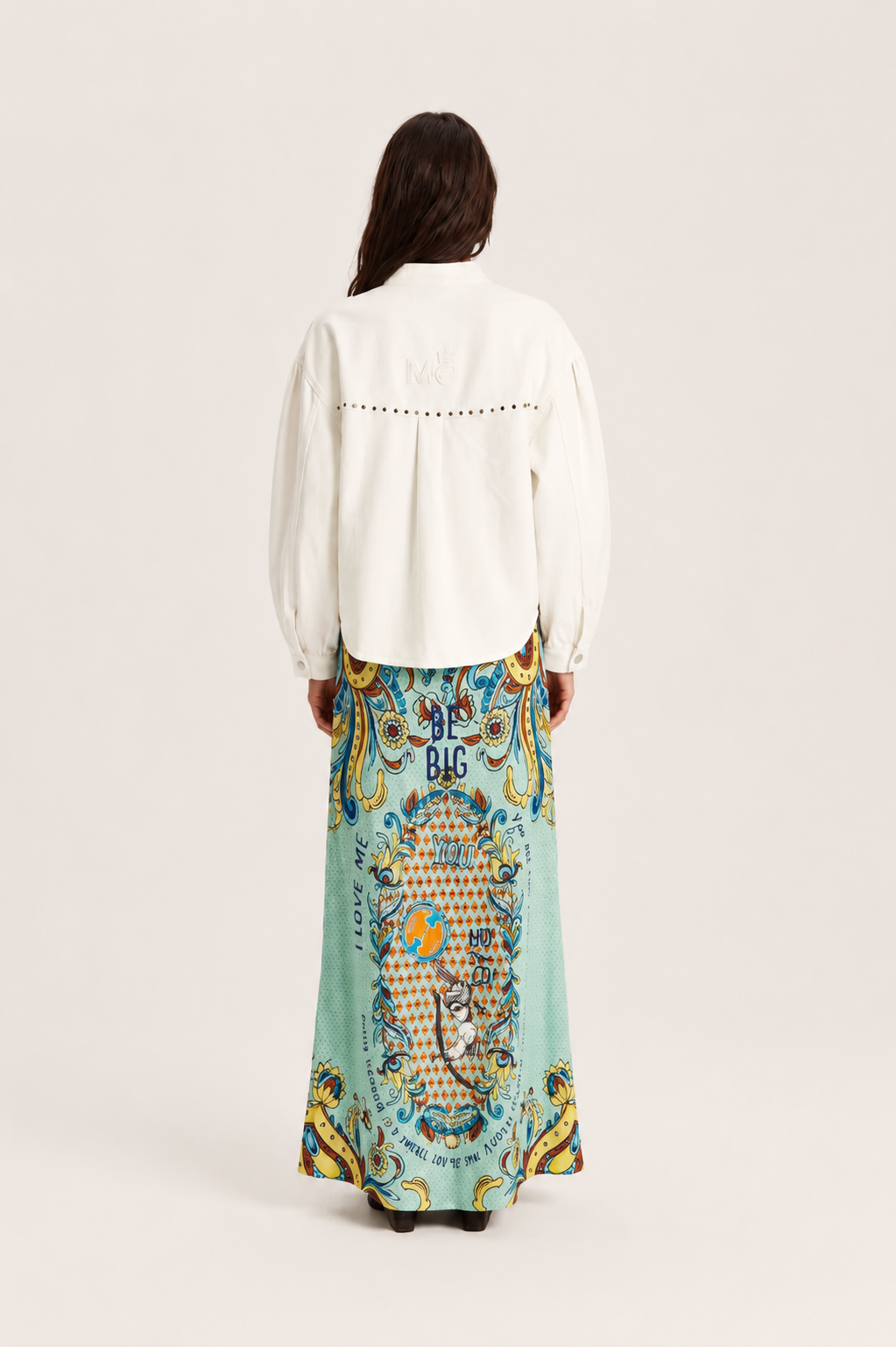 A woman shown from the back wearing a white long-sleeve blouse with decorative stitching and a long, wide skirt featuring a colorful baroque-style print in turquoise, gold, and blue.